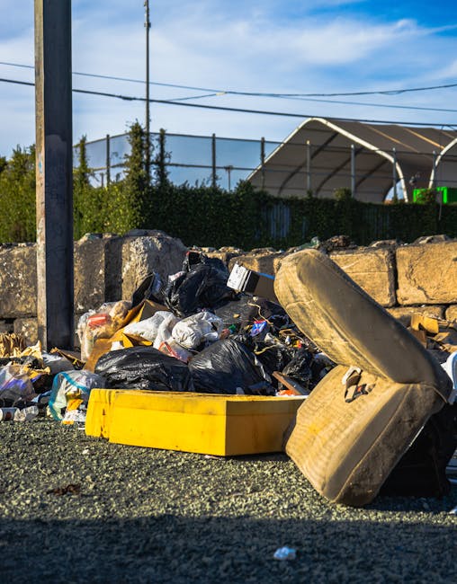 A visible accumulation of mixed waste spilling onto a paved surface in an outdoor setting, comprising black and clear plastic rubbish bags, discarded cardboard boxes, and a large, dirty, beige-colored car tire leaning against the pile. The waste materials appear haphazardly gathered, with some items partially covered by the bags and others loosely scattered. Behind the rubbish, there is a low stone wall constructed from roughly cut stones, partially obscuring the view of a chain-link fence topped with greenery and a structure with a peaked roof, possibly a sports facility or storage shed. A utility pole with multiple electrical wires extends vertically in the left side of the image, and the sky is partly cloudy with patches of blue, indicating daytime natural light. The scene offers an example of private waste disposal that could be handled by independent rubbish removal services like Rubbish Removal Holland Park, focusing on bulky rubbish collections and on-site clearance options for household or industrial waste, especially in urban or suburban environments.