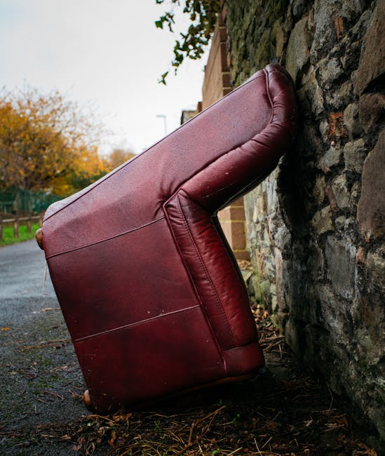 A large, red leather armchair with visible stitching and a plush padded backrest is positioned on a paved sidewalk, leaning against a rough-textured stone wall. The chair's upholstery shows some signs of wear, with slight creases and scratches, indicating it is used furniture being discarded. The armchair's legs are small and wooden, with a dark finish, and are partially visible on the uneven ground covered with scattered leaves and debris. The surrounding environment includes a brick building partially obscured by green foliage at the top of the image and a street extending into the distance, suggesting an outdoor urban or residential setting. Overcast lighting casts soft shadows, emphasizing the textures of the leather and stone. This scene reflects an instance of private waste disposal, where furniture is left outside for collection, aligning with the context of independent rubbish removal services under the relevant local guidelines.