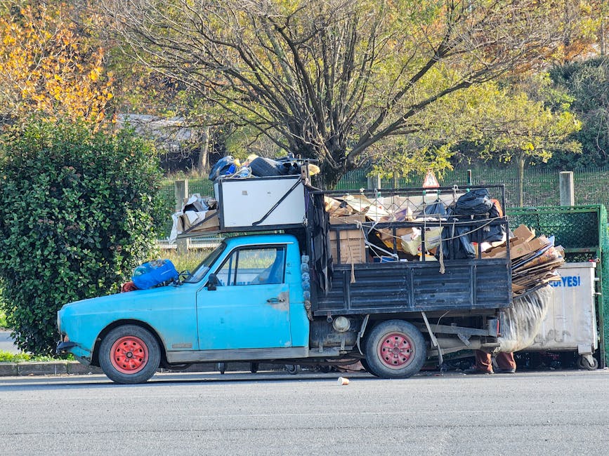 A visible accumulation of mixed waste spilling onto a paved surface in an outdoor setting, comprising black and clear plastic rubbish bags, discarded cardboard boxes, and a large, dirty, beige-colored car tire leaning against the pile. The waste materials appear haphazardly gathered, with some items partially covered by the bags and others loosely scattered. Behind the rubbish, there is a low stone wall constructed from roughly cut stones, partially obscuring the view of a chain-link fence topped with greenery and a structure with a peaked roof, possibly a sports facility or storage shed. A utility pole with multiple electrical wires extends vertically in the left side of the image, and the sky is partly cloudy with patches of blue, indicating daytime natural light. The scene offers an example of private waste disposal that could be handled by independent rubbish removal services like Rubbish Removal Holland Park, focusing on bulky rubbish collections and on-site clearance options for household or industrial waste, especially in urban or suburban environments.