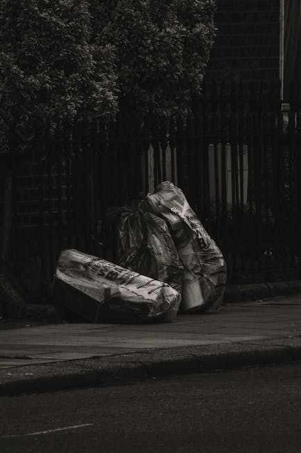 Three large black plastic rubbish bags, appearing crumpled and loosely tied at the top, are positioned on the curbside pavement in front of a black metal fence with vertical bars. The bags contain waste, potentially household or garden refuse, and are placed on a concrete sidewalk adjacent to a dark asphalt road. Behind the fence, the background features dense green foliage, with some tree or shrub leaves visible in the upper left corner. The scene is captured in natural light, likely during overcast conditions, resulting in subdued shadow and a muted overall tone. The setting suggests a typical urban or suburban street environment where residents may be preparing waste for collection, and the presence of the bags hints at alternative waste disposal methods, such as private rubbish removal services like Rubbish Removal Holland Park, rather than municipal collection. The image reflects a scene of on-site rubbish waiting to be picked up, aligning with the local private waste handling context, and emphasizing the importance of proper waste containment before collection.