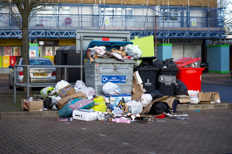 A large collection of mixed waste and rubbish is piled on the pavement in front of a park or commercial area, mainly consisting of cardboard boxes, plastic bags, paper, and several black trash bags. The waste includes flattened and unflattened cardboard boxes, some torn or crumpled, alongside loose paper and packaging materials scattered on the ground. A prominent grey recycling bin labeled for mixed paper and cardboard is positioned centrally, with its lid open, overflowing with various papers and parcels. To its right, black and red refuse bins are partially visible, with some trash spilling over nearby. In the background, there is a street scene featuring parked cars, including a grey vehicle with a visible license plate, and a building with a blue painted exterior and a commercial storefront, possibly a fish bar or takeaway, with closed metal shutters. A tree with a bare trunk is situated nearby, and a metal railing runs along the front, separating the waste area from the sidewalk, which comprises textured paving bricks. The environment appears to be during daytime with natural lighting. This scene illustrates common issues associated with improper rubbish disposal that can be addressed through private waste collection services like those offered by Rubbish Removal Holland Park, emphasizing the importance of correct waste management and than highlighting the potential need for efficient rubbish removal solutions in urban areas.