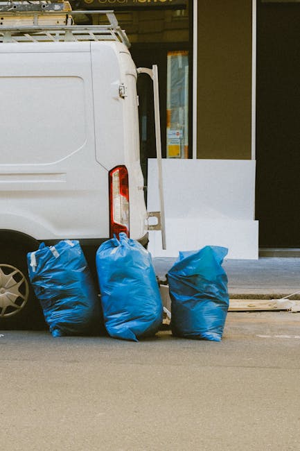 The image depicts the exterior of a residential building with green wooden cladding and red window frames, situated on a street with parked cars and a distant brick building in the background. At the base of the building, several black garbage bags are piled on the concrete pavement, some leaning against the corner of the structure. The bags are filled with waste materials and have a slightly glossy appearance, suggesting plastic content. A small section of weathered concrete steps leading to a white door with a window and a small overhanging roof is visible on the right side of the building. To the left, a graffiti-covered metal door with peeling paint and rust is partially shown. The surrounding environment includes a sidewalk, parking area, and urban infrastructure, with power lines visible overhead. The scene appears to capture a private waste collection or on-site rubbish removal scenario, managed independently rather than through local authority collection, reflecting common practices in urban waste management. Rubbish Removal Holland Park specialized in handling such waste disposal tasks, evident by the neatly gathered refuse outside the building, awaiting collection.