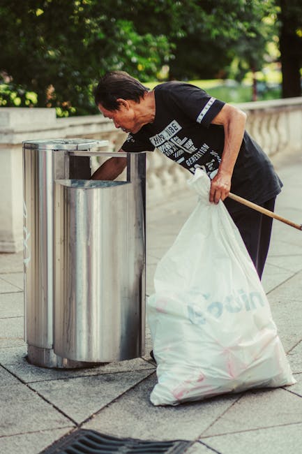 A man with dark hair, wearing a black T-shirt with white text and dark trousers, is seen bending over to dispose of trash into a cylindrical stainless steel public rubbish bin. He is holding a large white garbage bag, which appears to be filled with waste, in his left hand, while his right hand is reaching into the bin for placement. The bin has a smooth, reflective finish and is situated on a paved sidewalk that features textured stone tiles. In the background, there is a waist-high stone balustrade with a classic design, and lush green trees with bright foliage provide a natural backdrop, indicating a park or public space setting. The scene is well-lit with natural daylight, casting soft shadows, and the outdoor environment suggests an area where independent waste disposal or rubbish removal services might be employed as an alternative to council collection, consistent with the context of private rubbish handling in Holland Park.