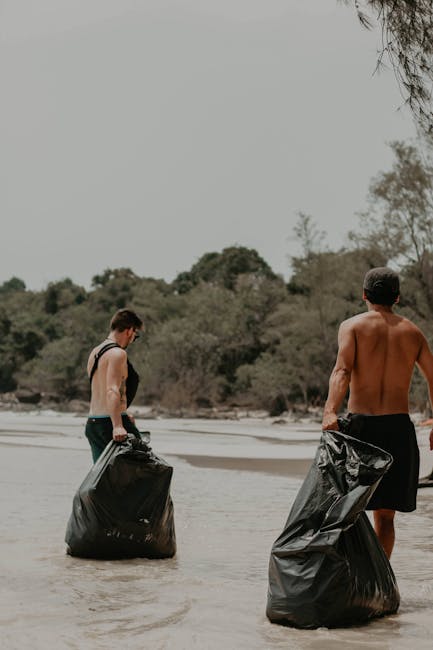 Two shirtless men are standing knee-deep in shallow water on a sandy beach, each holding large black rubbish bags filled with waste. The man on the left has short dark hair, is wearing dark shorts, and is looking down at his bag, which is partially submerged in the water. The man on the right, with short hair, is wearing dark shorts and is facing away from the camera, holding his rubbish bag in his right hand. The background features a natural environment with green trees, bushes, and rocky shoreline, under an overcast sky. The scene suggests an outdoor rubbish removal activity, possibly part of a beach clean-up effort, aligned with private or independent waste handling services like those offered by Rubbish Removal Holland Park, focusing on environmental responsibility and waste collection in natural settings.
