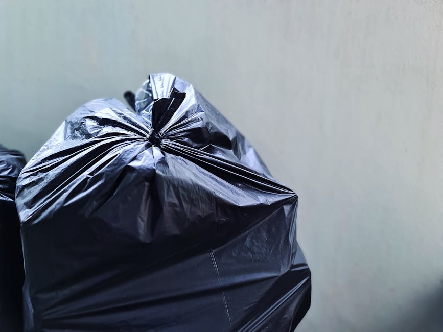 A man with dark hair, wearing a black T-shirt with white text and dark trousers, is seen bending over to dispose of trash into a cylindrical stainless steel public rubbish bin. He is holding a large white garbage bag, which appears to be filled with waste, in his left hand, while his right hand is reaching into the bin for placement. The bin has a smooth, reflective finish and is situated on a paved sidewalk that features textured stone tiles. In the background, there is a waist-high stone balustrade with a classic design, and lush green trees with bright foliage provide a natural backdrop, indicating a park or public space setting. The scene is well-lit with natural daylight, casting soft shadows, and the outdoor environment suggests an area where independent waste disposal or rubbish removal services might be employed as an alternative to council collection, consistent with the context of private rubbish handling in Holland Park.