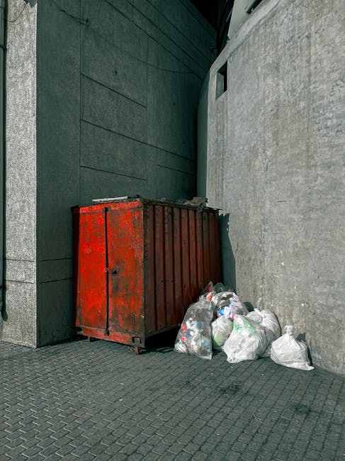 A red, weathered metal waste container, positioned against a dark green wall and a light grey concrete wall in an urban alleyway or narrow street setting, with its surface showing signs of rust and paint chipping. Several white plastic trash bags filled with mixed waste, including crumpled packaging and miscellaneous debris, are piled beside and partially leaning against the container. The ground is paved with dark, textured asphalt tiles. The environment appears tight and enclosed, with minimal natural light, suggesting this area is used for informal rubbish disposal or private waste clearance outside of official street collection routines. The presence of the waste container and bags indicates an alternative rubbish removal method, possibly handled independently by Rubbish Removal Holland Park, in line with their specialist services for difficult access rubbish jobs in residential or commercial mews areas.