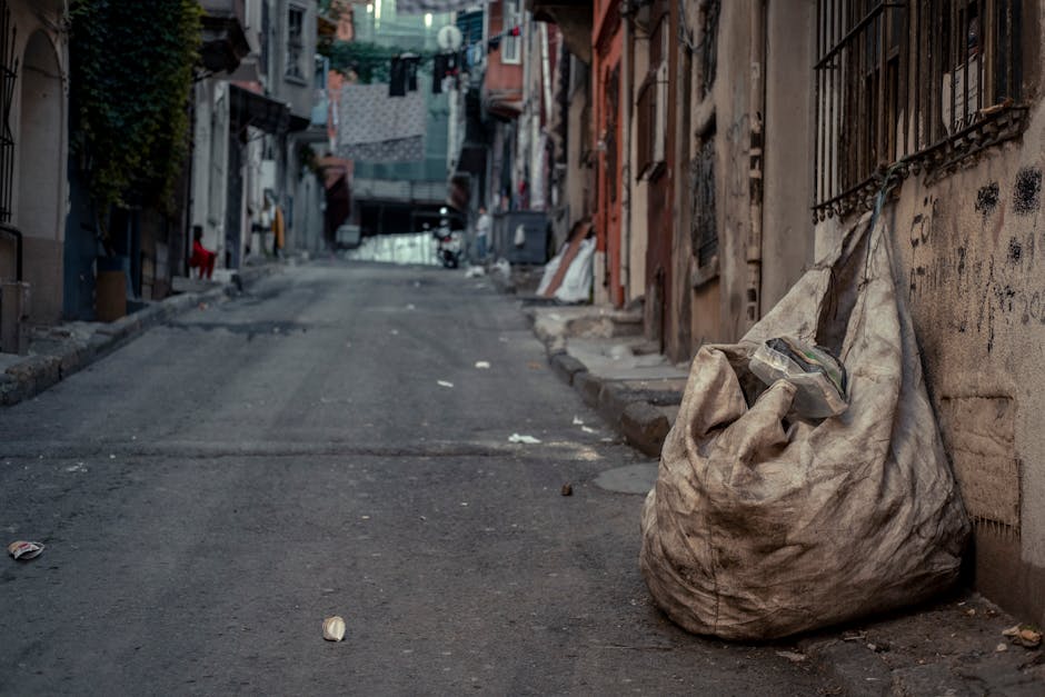 A middle-aged man with dark hair, dressed in a black T-shirt with white text and dark trousers, is seen disposing of rubbish into a cylindrical stainless steel public trash bin located on a paved sidewalk. The man is bent forward, using his left hand to hold a large, loosely tied white plastic bag filled with waste, and with his right hand, he is reaching into the opening of the bin. The background features lush green trees and foliage, creating a natural outdoor environment, with sunlight filtering through the leaves. The trash bin has a sleek, modern design with a rounded top opening, positioned near a low stone or concrete balustrade, indicating a public park or urban outdoor space. The scene suggests an act of private waste disposal, aligning with the services of rubbish removal specialists like Rubbish Removal Holland Park, who facilitate on-site collection and environmental responsibility in managing household or commercial rubbish.