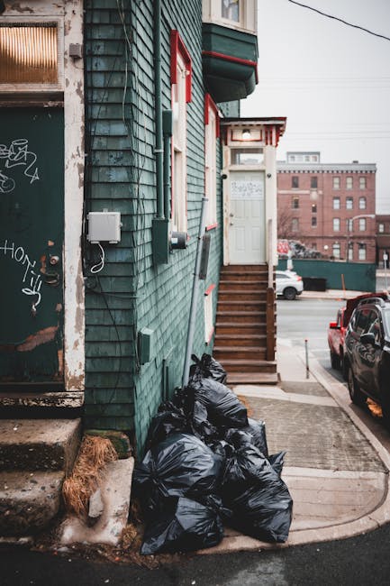 The image depicts a street scene featuring a building with teal-painted wooden cladding and red trim around the windows and door. A set of wooden stairs with a handrail leads up to a white door on the first floor of the building. On the concrete pavement beside the building, there are several tightly packed black refuse bags, filled with waste material, resting against the building's lower wall. To the left of the bags, part of a concrete step and a small patch of what appears to be snow or ice are visible. In the background, the street extends with a red car parked alongside, and other urban buildings, including a taller brick structure, are visible further down the road. The environment appears to be an inner-city area, with utility meters and cables attached to the building exterior, indicative of private waste disposal rather than council collection, consistent with private rubbish removal services like those provided by Rubbish Removal Holland Park. The overall scene conveys a typical urban waste presentation, emphasizing the need for timely rubbish clearance in a residential setting.