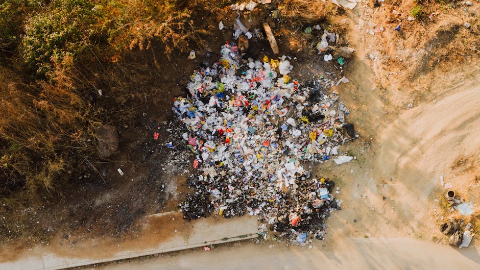 An aerial view of a large pile of mixed waste and household rubbish, including plastic bottles, bags, and packaging materials, scattered on bare soil at the edge of a dirt pathway. The pile is situated adjacent to a patch of dry, sparse vegetation, with some bushes visible in the upper left corner. The waste appears loosely accumulated with some items stacked and others spread out, indicating an informal or illegal dumping site. The soil around the rubbish shows signs of disturbance, with areas of darker, possibly burned material. In the lower part of the image, a curved section of a paved or concrete pathway runs alongside the scene. The environment suggests an outdoor location, potentially in a less maintained or neglected area where private waste disposal occurs without proper collection services, aligning with the context of alternative rubbish removal and illegal dumping issues. Rubbish Removal Holland Park often addresses such sites through careful on-site clearance to prevent environmental hazards.