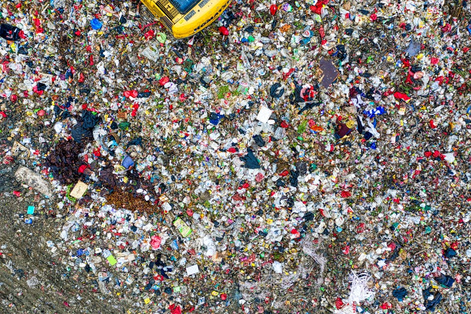 An aerial view of a large pile of mixed waste and household rubbish, including plastic bottles, bags, and packaging materials, scattered on bare soil at the edge of a dirt pathway. The pile is situated adjacent to a patch of dry, sparse vegetation, with some bushes visible in the upper left corner. The waste appears loosely accumulated with some items stacked and others spread out, indicating an informal or illegal dumping site. The soil around the rubbish shows signs of disturbance, with areas of darker, possibly burned material. In the lower part of the image, a curved section of a paved or concrete pathway runs alongside the scene. The environment suggests an outdoor location, potentially in a less maintained or neglected area where private waste disposal occurs without proper collection services, aligning with the context of alternative rubbish removal and illegal dumping issues. Rubbish Removal Holland Park often addresses such sites through careful on-site clearance to prevent environmental hazards.