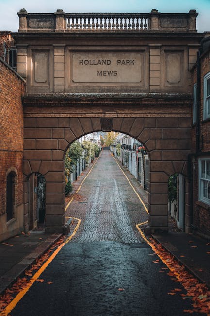 The image shows a narrow, slightly inclined cobblestone street viewed through a large stone archway, which is part of a decorative stone bridge structure with classical detailing and a landscaped balustrade on top. The bridge bears the inscription 'HOLLAND PARK MEWS' carved into a rectangular stone panel. On either side of the street, there are residential buildings with brick and stone facades, some with bay windows and decorative window surrounds. The street is lined with narrow pavement pathways and appears to be in a quiet, residential area. The scene is captured during an overcast day, with soft, diffuse lighting, and fallen autumn leaves scattered along the edges of the street. The overall environment suggests a typical London suburb, where private waste collection or alternative rubbish removal services, such as that provided by Rubbish Removal Holland Park, may operate to maintain the cleanliness of these historic streetscape and terraced dwellings.