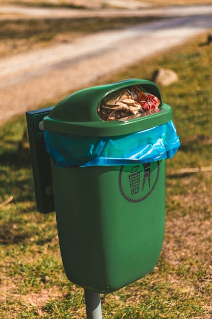 A green outdoor rubbish bin with a semi-transparent, slightly domed lid filled with assorted waste, including crumpled paper and plastic wrappers. The bin is mounted on a metal pole and positioned on a grassy area near a gravel pathway. A blue plastic liner is visible inside the bin, secured around the rim. The environment in the background includes a dirt pathway and blurred natural scenery, suggesting a park or open public space. The bin features a black graphic symbol of a trash can with a line through it, indicating proper waste disposal, reflecting efforts to promote cleanliness and alternative waste handling solutions, such as private collection services like Rubbish Removal Holland Park.