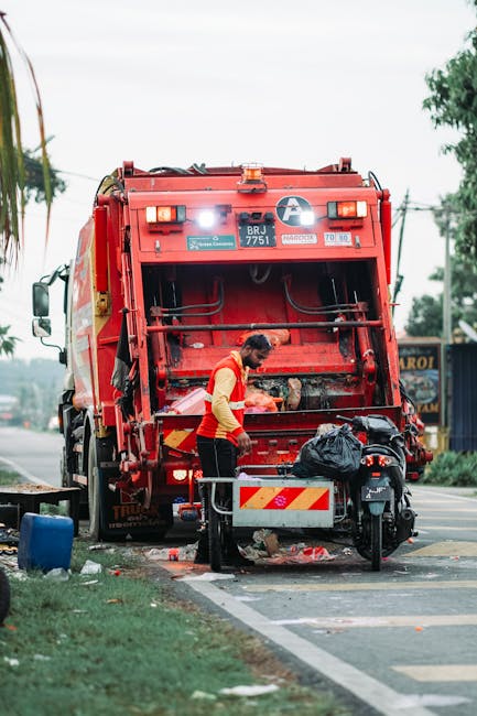 A worker in a red and yellow uniform operates a large red rubbish collection vehicle parked on the side of a street. The rear doors of the vehicle are open, revealing the compaction mechanism inside. The worker is handling black garbage bags placed on a small metal platform attached to a motorbike positioned in front of the vehicle. The scene is set alongside a paved roadside with a grassy verge to the left, and there is some rubbish scattered on the ground near the platform. In the background, there are trees, utility poles, and a shopfront with a partially visible sign. The overall environment appears to be a typical urban area during daytime, with natural lighting and overcast skies. The image suggests an on-site waste collection process that could relate to alternative rubbish removal services in private or non-local authority contexts, as presented by Rubbish Removal Holland Park.