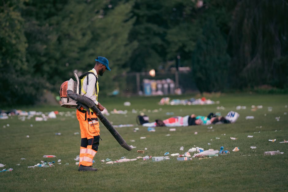 A professional worker wearing high-visibility orange trousers with reflective stripes, a white jacket, and a blue cap is seen using a leaf blower attachment on a hose to clear litter from a grassy park area. The worker is positioned in the foreground, focused on cleaning up the scattered debris, which includes plastic bottles, cups, and paper waste spread across the grass. In the background, there are trees with dense foliage and a group of collapsed individuals lying on the ground, possibly resting or unconscious, amidst more scattered rubbish. To the far back, a row of trash containers and some outdoor structures are visible, indicating a managed park environment. The lighting suggests late afternoon or early evening, with a natural outdoor setting. This scene illustrates the importance of waste removal and environmental cleaning services that could be provided by companies like Rubbish Removal Holland Park as part of alternative waste handling methods to maintain cleanliness near public parks.