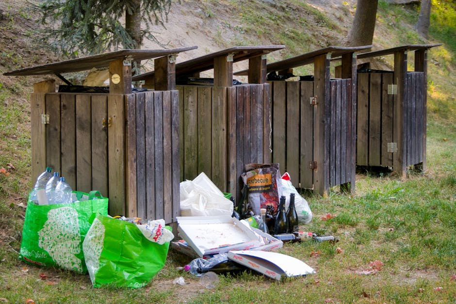 An outdoor scene depicts four wooden waste bins positioned in a row on a grassy area beside a sloped, wooded background with trees and foliage. The bins are made of vertical wooden planks stained in dark brown, with partially open lids, some slightly tilted. In front of the leftmost bin, there are several green reusable shopping bags filled with plastic bottles and other waste, alongside a white pizza box with remnants of food. To the right, there are additional discarded items including a cardboard box labeled 'barbecue,' a few glass bottles, plastic bags, and miscellaneous refuse scattered on the ground. The environment is illuminated by natural daylight, highlighting the textures of the wood and the various waste materials, which seem to be left for either alternative rubbish disposal or private collection. The scene reflects an example of illegal dumping or unmanaged rubbish, illustrating the need for proper waste removal services such as those provided by Rubbish Removal Holland Park.